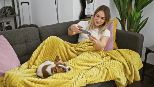 Μια χαμογελαστή νεαρή γυναίκα τραβάει φωτογραφίες του χαριτωμένου σκύλου της, ενώ χαλαρώνει σε μια κίτρινη κουβέρτα σε έναν καναπέ στο σπίτι. - Πλάνα, βίντεο