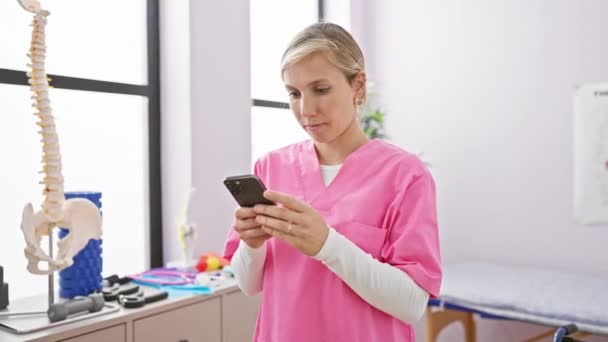 Una donna concentrata in scrub rosa utilizza uno smartphone in una clinica di riabilitazione ben illuminata. - Filmati, video