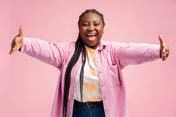 Belle, souriante excitée corps positif femme afro-américaine avec étreinte regardant la caméra. Jeune femelle isolée sur fond rose. Concept de bienvenue - Photo, image