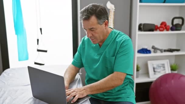 Un uomo maturo in scrub che usa un portatile nella stanza di una moderna clinica di riabilitazione. - Filmati, video