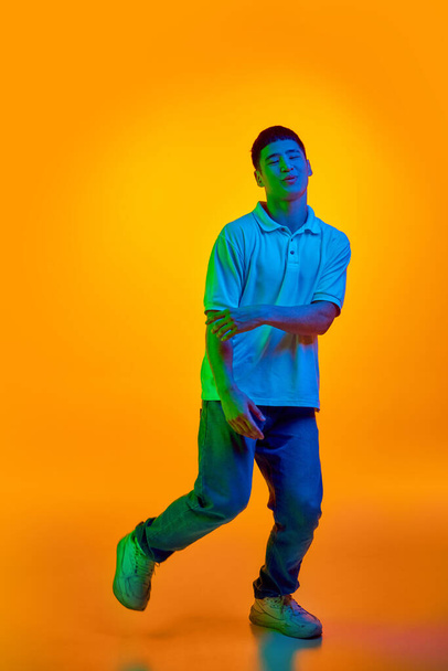 Teljes test fotó vicces, vidám, fiatal ázsiai férfi táncol örömmel mozgó élénk neon fény ellen gradiens narancs háttér. Az emberi érzelmek, divat és szépség, munka és hobbi fogalma. - Fotó, kép