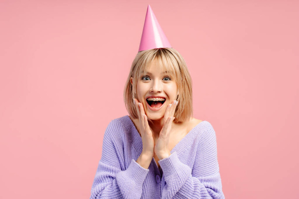 Mutlu, heyecanlı bir kadın, parti şapkası takmış, pembe arka planda izole edilmiş doğum gününü kutluyor. Tatil kavramı  - Fotoğraf, Görsel