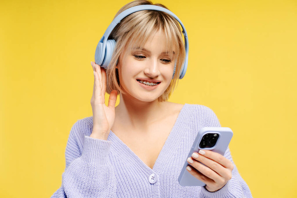 Portret van een lachende leuke tienervrouw met een koptelefoon, een mobiele telefoon in haar hand, luisterend naar muziek geïsoleerd op een gele achtergrond. Leuk concept, technologie  - Foto, afbeelding