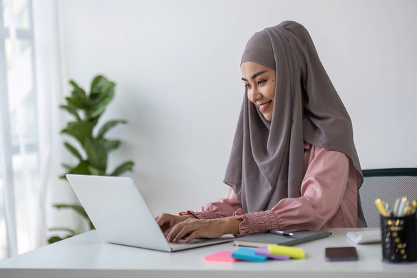 Lächelnde muslimische Geschäftsfrau, die einen Hidschab trägt, während sie am Schreibtisch im Büro online chattet.. - Foto, Bild