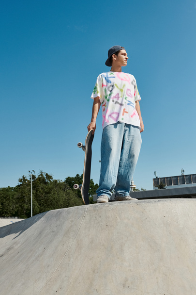 Ένα νεαρό αγόρι σκέιτερ με αυτοπεποίθηση στέκεται στην κορυφή μιας ράμπας skateboard, έτοιμο να εκτελέσει τολμηρά κόλπα σε ένα καλοκαιρινό πάρκο skate. - Φωτογραφία, εικόνα