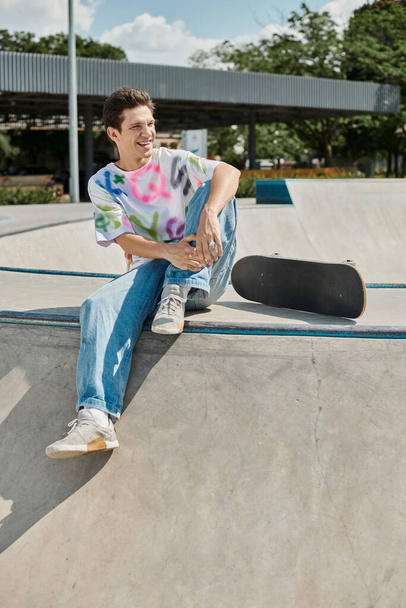 Een jongeman zit gepensioneerd aan de rand van een skateboard helling, te genieten van de sensatie van de naderende afdaling in een skate park. - Foto, afbeelding