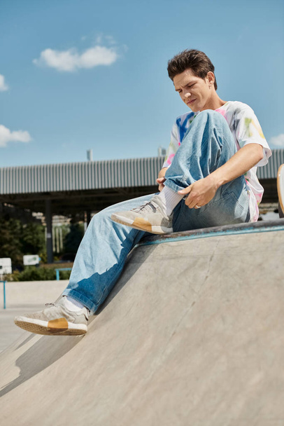 Ένας νεαρός σκέιτερ με αυτοπεποίθηση κάθεται στην κορυφή μιας ράμπας skateboard σε ένα ζωντανό υπαίθριο πάρκο skate σε μια ηλιόλουστη μέρα του καλοκαιριού. - Φωτογραφία, εικόνα