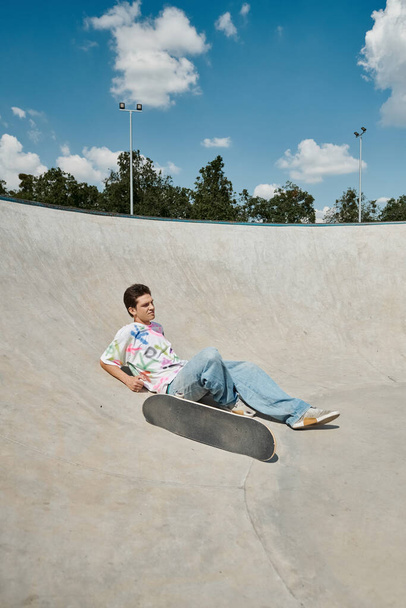 Egy fiatal korcsolyázó fiú a gördeszka mellett pihen egy rámpán egy nyüzsgő szabadtéri gördeszka parkban egy napos nyári napon.. - Fotó, kép