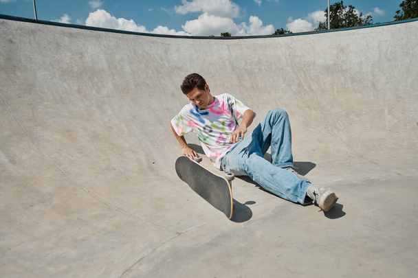 Ένα νεαρό αγόρι σκέιτερ αψηφά τη βαρύτητα, κάθεται κοντά skateboard στη ράμπα σε ένα υπαίθριο πάρκο skate σε μια ηλιόλουστη μέρα του καλοκαιριού. - Φωτογραφία, εικόνα