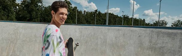 Un hombre joven adornado con una camisa vibrante sostiene alegremente un monopatín en un bullicioso parque de skate en un día soleado. - Foto, imagen