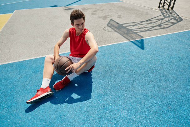 Egy fiatalember ül a kosárpályán, mélyen a gondolataiban, kosárlabdát tart egy napsütéses nyári napon.. - Fotó, kép