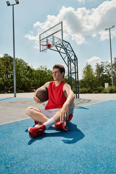 Mladý basketbalista sedí na hřišti, hluboce zamyšlený, drží v rukou basketbal za slunečného letního dne. - Fotografie, Obrázek