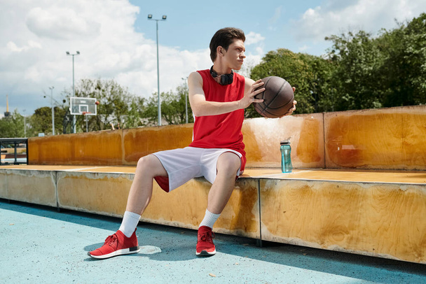 Ένας νεαρός άνδρας κάθεται σε ένα παγκάκι, βαθιά στη σκέψη, κρατώντας ένα μπάσκετ σε μια ηλιόλουστη μέρα. - Φωτογραφία, εικόνα