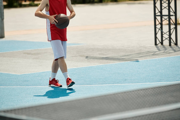 Ένας νεαρός άνδρας στέκεται σε ένα γήπεδο μπάσκετ κρατώντας μια μπάλα, έτοιμος να παίξει κάτω από τον καλοκαιρινό ήλιο. - Φωτογραφία, εικόνα
