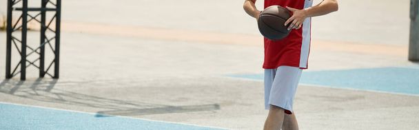 Egy fiatal kosaras áll a kosárlabda pálya tetején, magabiztosan tartja a labdát egy napos nyári napon.. - Fotó, kép