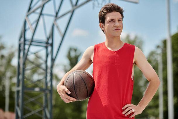Ένας νεαρός άνδρας με ένα ζωηρό κόκκινο πουκάμισο κρατά επιδέξια ένα μπάσκετ, ενώ παίζει έξω σε μια καλοκαιρινή μέρα. - Φωτογραφία, εικόνα