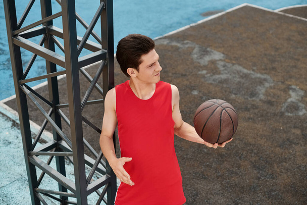 Młody mężczyzna ubrany na czerwono prezentuje swoje umiejętności obsługi koszykówki na świeżym powietrzu w letni dzień. - Zdjęcie, obraz