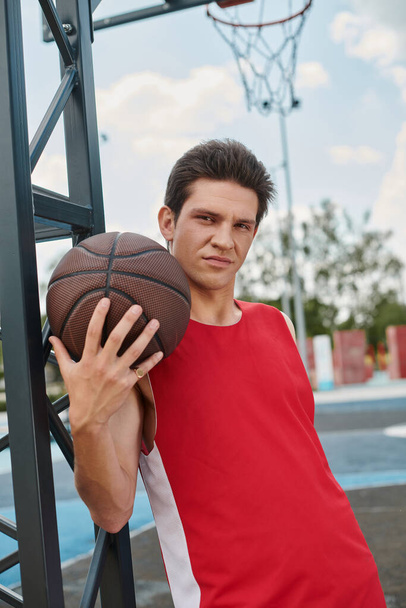 Молодой человек в красной рубашке умело держит баскетбол, готовый играть на открытом воздухе в летний день. - Фото, изображение