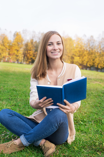 Studentin sitzt mit aufgeschlagenem blauen Buch auf grünem Gras - Foto, Bild