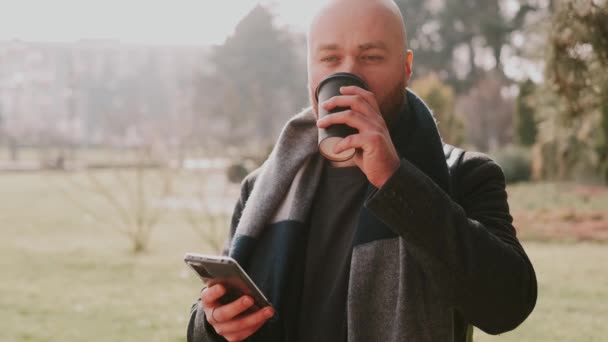 Záběry mladého vousatého muže pomocí smartphonu a pití šálek kávy jít ven v parku. - Záběry, video