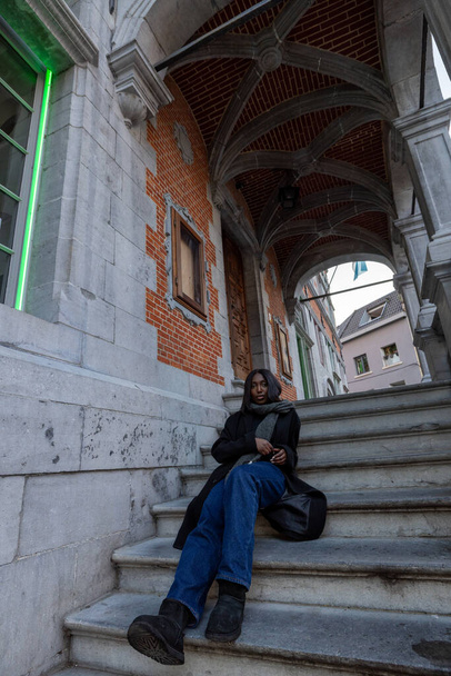 Esta imagen captura a una joven en un momento de reposo en los escalones de un edificio histórico. Los detalles arquitectónicos, incluyendo el pasillo arqueado y la obra de ladrillo, proporcionan una sensación clásica europea - Foto, imagen