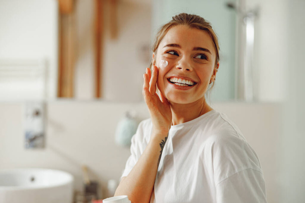 Mujer feliz aplicando hidratante hidratante en su cara sentada en el baño. Rutina de belleza - Foto, Imagen