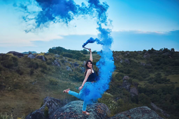 Piękna młoda kobieta w przyrodzie z niebieskim dymem w powietrzu. Światło i cień tworzą dramatyczny efekt. Osoba trzyma niebieską bombę dymną.. - Zdjęcie, obraz