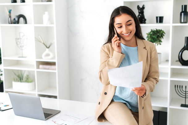 Успешная многорасовая женщина, сотрудник компании, генеральный директор, сидя на столе в офисе, разговаривая по смартфону, беседуя с партнером или клиентом, изучая финансовые документы, улыбаясь - Фото, изображение