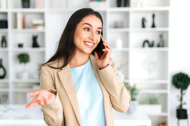 Беспроводная связь. Позитивная великолепная арабская или индийская женщина, руководитель компании, менеджер, беседующий по телефону с партнером или клиентом, обсуждающий вопросы работы, назначает встречу, улыбается - Фото, изображение