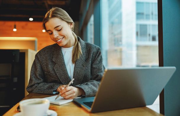 Bella donna d'affari prendendo appunti e navigando laptop seduto in coworking moderno - Foto, immagini