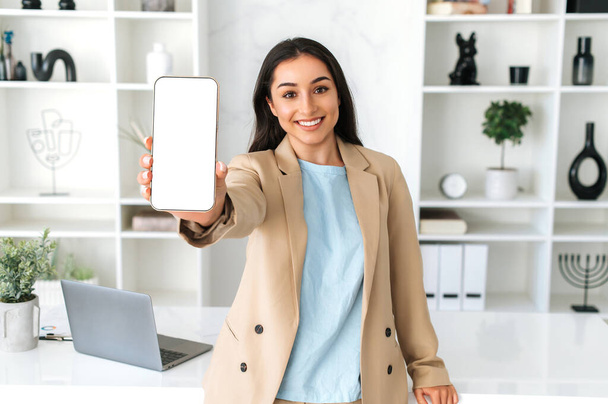 Копирование пространства, макет. Положительный довольно индийская или арабская женщина, сотрудник компании, стоять в офисе, показывая смартфон с чистым белым макетом экрана для рекламы или презентации, улыбка на камеру - Фото, изображение