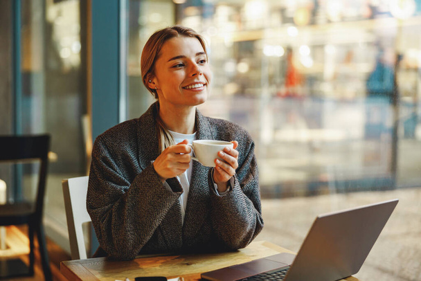 Улыбающаяся женщина-предприниматель пьет кофе в коворкинге во время работы на ноутбуке возле окна - Фото, изображение