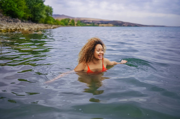 Приваблива щаслива плюс розмір афроамериканська чорна жінка насолоджується плаванням, розважаючись з бризками води в морському озері вода в хмарну погоду на відкритому повітрі - Фото, зображення