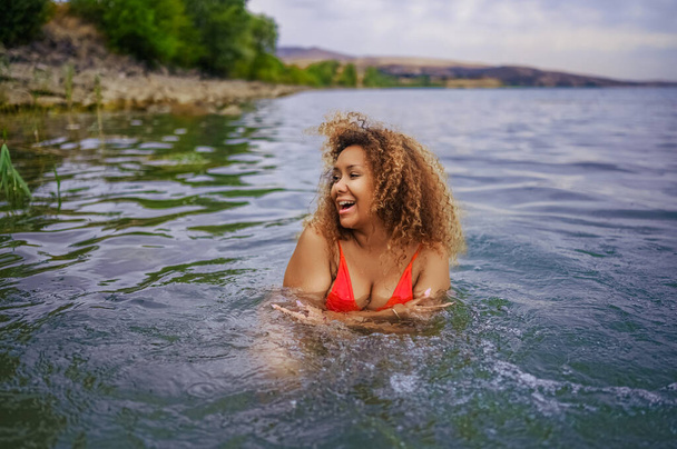 魅力的な幸せプラスサイズアフリカ系アメリカ人黒人女性は、曇った天候の屋外で海の湖の水で水のスプラッシュと一緒に水泳を楽しんでいます - 写真・画像