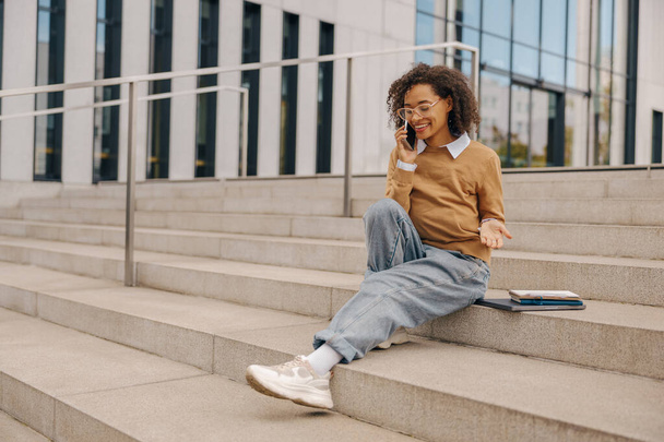 Sorridente telefono femminile freelance parlando con il cliente mentre seduto su sfondo edificio - Foto, immagini