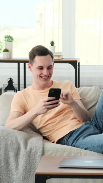 Radosny biały mężczyzna, który prowadzi ożywione rozmowy i wybucha śmiechem, korzystając z telefonu na kanapie. Z jasnym uśmiechem na twarzy i niepowtarzalnym błyskiem w oczach. - Materiał filmowy, wideo