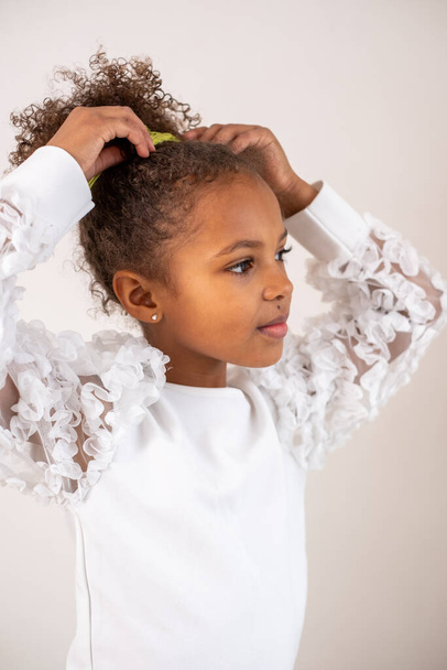 Une jeune fille métissée contemplative aux cheveux bouclés, vêtue d'une robe blanche à volants, regarde sur le côté sur un fond neutre, évoquant curiosité et élégance. Photo de haute qualité - Photo, image
