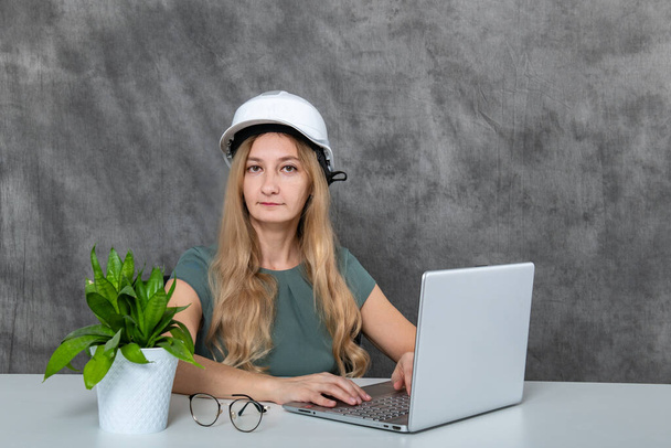 Μια νεαρή γυναίκα με ξανθά μαλλιά κάθεται και φοράει λευκό κράνος. Είναι συγκεντρωμένη και δακτυλογραφεί σε έναν φορητό υπολογιστή, δείχνοντας αφοσίωση και συγκέντρωση στη δουλειά της. - Φωτογραφία, εικόνα