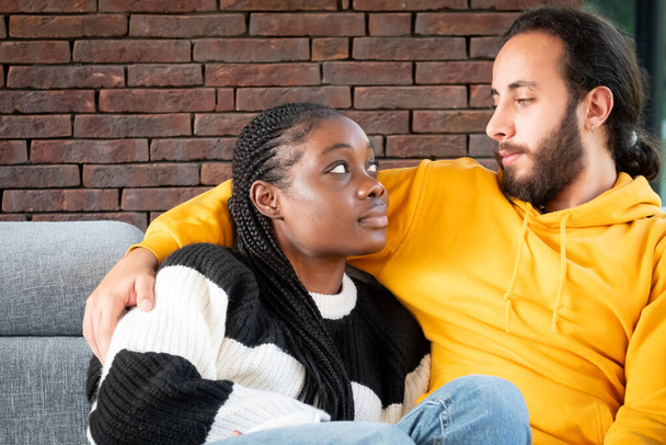 Dieses Bild zeigt einen zärtlichen und liebevollen Moment zwischen einem Paar in der Gemütlichkeit eines Hauses. Eine afroamerikanische Frau mit geflochtenen Haaren blickt zu ihrem Partner auf, einem Mann, der von mittlerer Statur sein könnte. - Foto, Bild