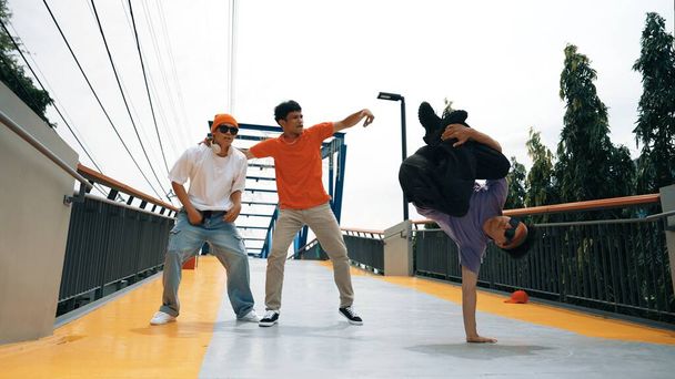 Панорамный снимок хипстера, одетого в стильную ткань и стоящего на месте. Группа профессиональных танцоров исполняет брейк-данс, в то время как выдающийся мужчина практикует брейк-бой. Открытый спорт 2024. Ошибочно - Фото, изображение