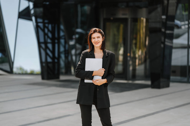 Cheerful young successful woman wearing suit carrying a laptop on a sunny day near a business building. - Photo, Image