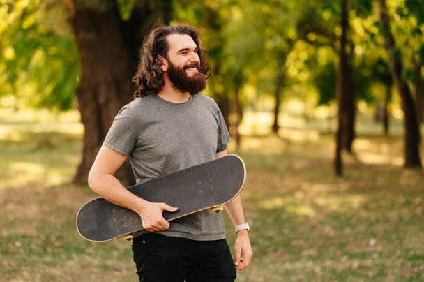 Uzun saçlı, gülümseyen sakallı adam parkta yürürken kaykayını tutuyor.. - Fotoğraf, Görsel