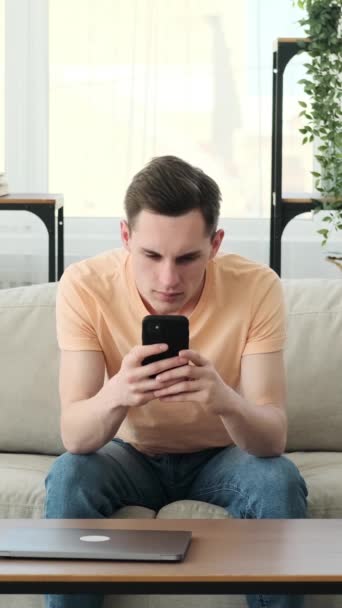 Man gaat in gesprek via zijn telefoon in het comfort van zijn huis. Deze jongeman, terloops gekleed, vindt zichzelf boeiend berichten zijn scherm, zijn wenkbrauwen gefronst in verbazing. - Video