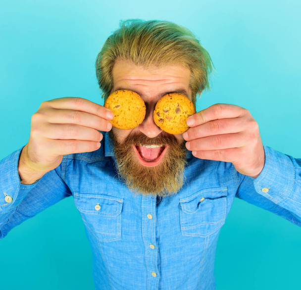 Bearded man covered eyes with freshly baked oatmeal cookies. Tasty sweet snack. Handsome male eating chocolate cookies biscuit. Breakfast or lunch. Smiling guy with delicious homemade cookies. Bakery - Photo, Image