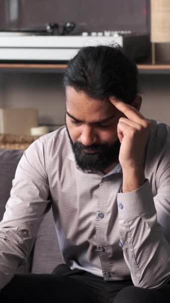 Trabajador de oficina indio, sufre un dolor de cabeza mientras descansa en un sofá en la sala de estar. Las expresiones transmiten los desafíos y tensiones de la jornada laboral, incluso en el entorno familiar del hogar. - Metraje, vídeo