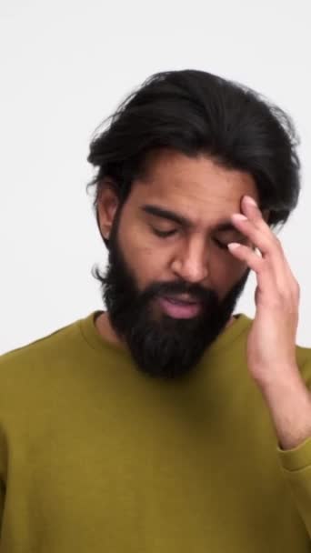 Kimerült szakállas fiatalember eltávolítja a szemüveget, és dörzsöli a fejét a fájdalom, miközben áll elszigetelt fehér háttér - Felvétel, videó