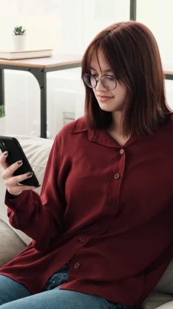Een Kaukasisch tienermeisje, zittend op een bank, is bezig met het gebruik van de telefoon. Met gefocuste aandacht navigeert ze door apps, berichten en sociale media, met een mix van nieuwsgierigheid en betrokkenheid. - Video