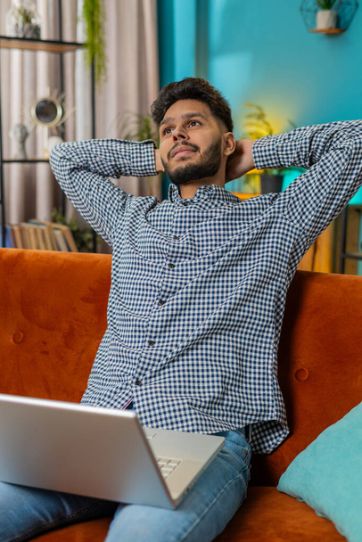 Indián férfi ül a kanapén, laptopot zárva, miután befejezte a munkát a nappaliban. Arab srác szabadúszó dolgozik online távoli munkát otthon kanapé. E-learning böngészés internet notebook számítógépen. Függőleges - Fotó, kép