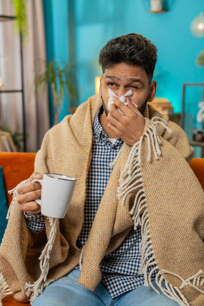 Intialainen sairas mies kärsii kylmästä tai allergiasta makaa sohvalla olohuoneessa kotona. Sairas latino kaveri pyyhe päässä puhaltaa aivastuksia pyyhkii räkää lautasliinaan. Coronavirus karanteeni pandemia. Pystysuora - Valokuva, kuva