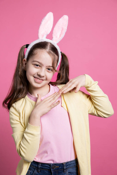Roztomilá mladá dívka tančí a pohrává si s kamerou, má na sobě králičí uši a copánky přes růžové pozadí. Bezstarostná školačka pocit radosti a nadšení z velikonoční oslavy ve studiu. - Fotografie, Obrázek